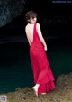 A woman in a red dress standing on a rock by the water.