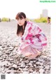 A woman in a pink and white plaid dress crouching on a rocky beach.